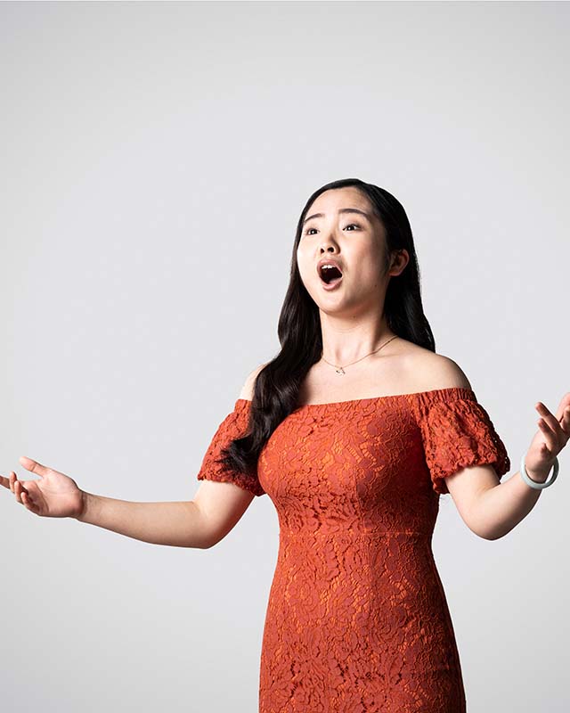 An RCM student in a red dress singing, against a grey background